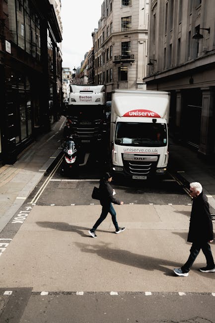 A street scene in an urban area with multi-storey buildings on both sides, featuring a small flatbed truck and a larger moving van parked parallel to the sidewalk, both displaying the logo 'Uniserve' and website 'uniserve.co.uk.' A motorcycle is parked in front of the vehicles, partially visible. The street has a designated loading zone marked with white lines and a sign, with the pavement level and curb visible. Two pedestrians are crossing the road: a woman wearing dark clothing and carrying a backpack, and an older man in a black coat. The overcast sky casts diffuse light over the scene. The setting suggests a home relocation or furniture transport process, with the vehicles ready for loading or unloading items as part of a professional moving service by Man and Van Kentish Town.