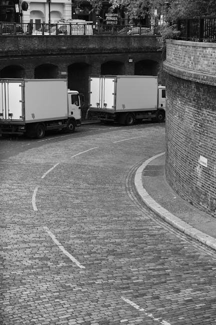 A black-and-white photograph shows two medium-sized commercial boxes trucks parked on a narrow, curved cobblestone street surrounded by brick walls and metal railings. The trucks are positioned side by side, facing away from the viewer, with their rear doors closed. The street has painted white dashed lines indicating parking or loading zones and is slightly inclined, leading towards an underpass or bridge supported by concrete pillars in the background. Above the brick wall, there is a fence and some trees visible, suggesting an urban residential area. The environment appears quiet, with no visible pedestrians or other vehicles. This scene reflects elements of home relocation or furniture transport logistics, as trucks are prepared for loading or unloading. During a house move or removal service by Man and Van Kentish Town, such vehicles are often used to transport belongings, furniture, and boxes from properties to new locations, fitting the context of packing and moving activities related to the webpage avoid hidden removals charges in Kentish Town NW5.