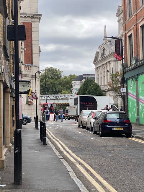 A street scene in an urban area with multi-storey buildings on both sides, featuring a small flatbed truck and a larger moving van parked parallel to the sidewalk, both displaying the logo 'Uniserve' and website 'uniserve.co.uk.' A motorcycle is parked in front of the vehicles, partially visible. The street has a designated loading zone marked with white lines and a sign, with the pavement level and curb visible. Two pedestrians are crossing the road: a woman wearing dark clothing and carrying a backpack, and an older man in a black coat. The overcast sky casts diffuse light over the scene. The setting suggests a home relocation or furniture transport process, with the vehicles ready for loading or unloading items as part of a professional moving service by Man and Van Kentish Town.