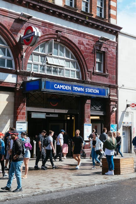 Outside Camden Town Station in Kentish Town, a group of people are gathered on the pavement, some standing and others walking towards or away from the station entrance. The station's brick facade features arched windows and a large blue sign reading 'CAMDEN TOWN STATION' positioned above the entrance, with the iconic London Underground roundel logo visible nearby. Several individuals are carrying backpacks and suitcases, suggesting they may be involved in a home relocation or moving process. In the foreground, a wooden planter box is placed on the pavement, and the scene is lit by daylight, reflecting a typical busy day in an urban environment. The context relates to a relocation or moving service, such as those offered by Man and Van Kentish Town, connected to parking regulations and logistical considerations for house removals in the area.