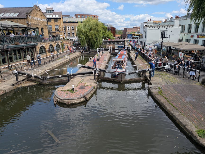 A canal scene in a lively urban area showing a narrowboat moored in the water, with a small bridge for pedestrians crossing over the canal. On one side, there is a paved walkway with several people walking and gathering, some observing the canal, and others engaged in activities related to house removals such as lifting or carrying boxes. Adjacent to the canal, a multi-story building with outdoor seating and a balcony is visible, with people sitting at tables. The building is constructed with brick and has large windows. On the opposite side, there are shops and cafes storefronts with outdoor tables and chairs, some occupied by patrons. A number of cardboard boxes, plastic-wrapped items, and furniture parts are near the area, indicating packing or move-in activities. The scene is well-lit with natural daylight, and a large green tree provides some shade. This setting likely depicts a community area suitable for home relocation logistics, with [COMPANY_NAME] occasionally involved in furniture transport or packing during the moving process.