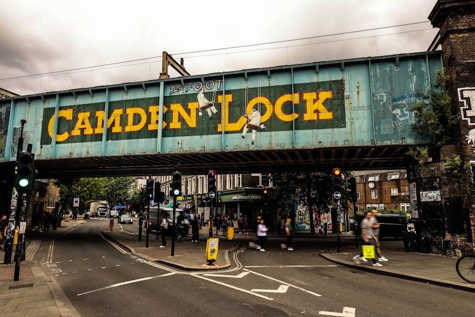 Outside Camden Town Station in Kentish Town, a group of people are gathered on the pavement, some standing and others walking towards or away from the station entrance. The station's brick facade features arched windows and a large blue sign reading 'CAMDEN TOWN STATION' positioned above the entrance, with the iconic London Underground roundel logo visible nearby. Several individuals are carrying backpacks and suitcases, suggesting they may be involved in a home relocation or moving process. In the foreground, a wooden planter box is placed on the pavement, and the scene is lit by daylight, reflecting a typical busy day in an urban environment. The context relates to a relocation or moving service, such as those offered by Man and Van Kentish Town, connected to parking regulations and logistical considerations for house removals in the area.