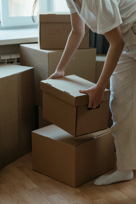 A person wearing a beige outfit is shown stacking plain brown cardboard moving boxes of various sizes on a wooden floor inside a well-lit room. The individual is holding a box with both hands, preparing for a home relocation process. Additional boxes are placed on the floor nearby, and one with the handwritten label 'CLOTH' is visible on top of a larger box. The setting appears to be an interior space, possibly a living room or storage area, with natural light coming through a nearby window. This scene illustrates the packing stage of furniture transport and moving logistics, which is part of house removals services offered by Man and Van Kentish Town, aligned with professional packing and loading processes.