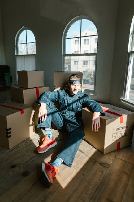 A person wearing a beige outfit is shown stacking plain brown cardboard moving boxes of various sizes on a wooden floor inside a well-lit room. The individual is holding a box with both hands, preparing for a home relocation process. Additional boxes are placed on the floor nearby, and one with the handwritten label 'CLOTH' is visible on top of a larger box. The setting appears to be an interior space, possibly a living room or storage area, with natural light coming through a nearby window. This scene illustrates the packing stage of furniture transport and moving logistics, which is part of house removals services offered by Man and Van Kentish Town, aligned with professional packing and loading processes.