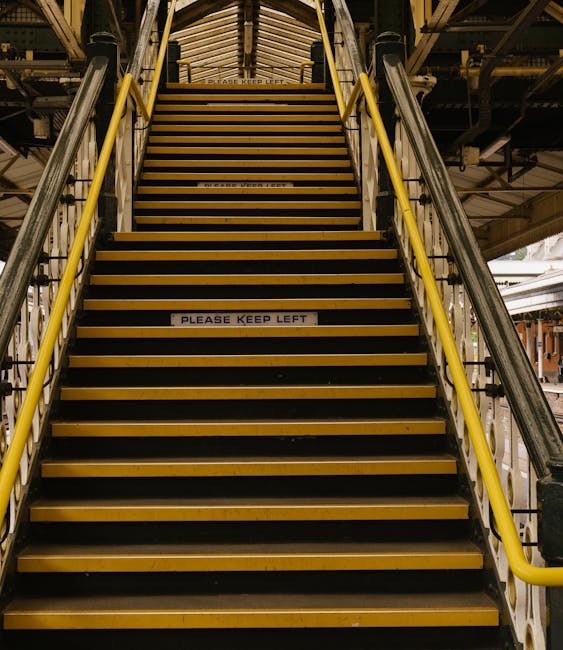A wide, indoor staircase with black anti-slip treads and yellow safety handrails on both sides, leading up to an upper level in a warehouse or industrial building. The stairs are constructed of metal, with a wooden ceiling visible at the top. There are two signs on the stairs reading 'PLEASE KEEP LEFT' in black text on white background, indicating traffic flow during moving or loading operations. The surroundings include metal structural components, pipes, and lighting fixtures, suggesting an active environment used for house removals or furniture transport, supported by the context of vehicle loading areas nearby. The overall scene captures a moment where furniture and boxes might be delivered or moved, consistent with home relocation services offered by companies like Man and Van Kentish Town.