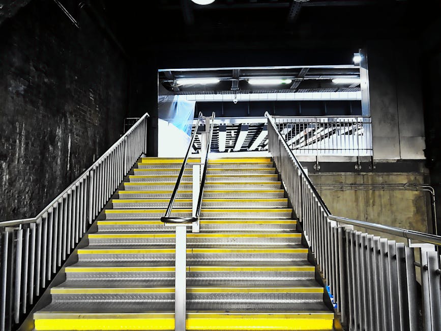 A wide, indoor staircase with black anti-slip treads and yellow safety handrails on both sides, leading up to an upper level in a warehouse or industrial building. The stairs are constructed of metal, with a wooden ceiling visible at the top. There are two signs on the stairs reading 'PLEASE KEEP LEFT' in black text on white background, indicating traffic flow during moving or loading operations. The surroundings include metal structural components, pipes, and lighting fixtures, suggesting an active environment used for house removals or furniture transport, supported by the context of vehicle loading areas nearby. The overall scene captures a moment where furniture and boxes might be delivered or moved, consistent with home relocation services offered by companies like Man and Van Kentish Town.