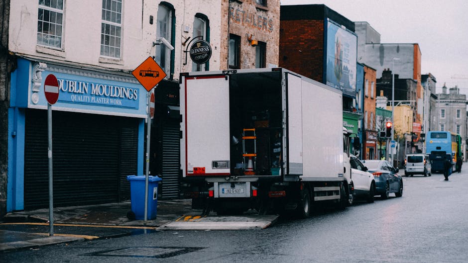 A white moving truck with an open rear door is parked on an urban street outside a row of shops in Kentish Town, with several cardboard boxes and wrapped furniture inside. The truck is positioned beside the pavement, partially on a loading ramp, with a forklift extension visible near the open garage. Surrounding the truck are parked cars, and shopfronts with signage, including a blue storefront labeled 'DUBLIN MOULDINGS' and a pub sign for 'GUINNESS,' indicating a commercial area. Safety and traffic signs, such as a no-entry sign and a warning sign for roadwork, are mounted on poles nearby. The scene appears to capture the loading process involved in a home relocation or furniture transport, with storage materials like cardboard boxes and plastic wrap visible in the vehicle, set against overcast lighting typical of an overcast day in Kentish Town, supporting the context of professional removals.