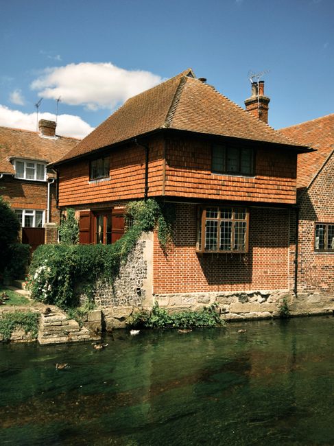 A traditional brick house with a steeply pitched tiled roof situated beside a waterway, with a small stone retaining wall along the water's edge. The house features multiple windows, including a prominent bay window on the ground floor and smaller windows on the upper level. There is greenery, such as climbing plants and shrubs, growing along the lower part of the building, which adds to the exterior's charm. The surrounding area includes other residential buildings with similar roofing styles, visible under a partly cloudy sky. The image was taken outdoors during daylight, capturing the house’s exterior and immediate environment. This setting reflects typical residential properties involved in house removals or relocation services, with the house possibly being prepared for packing, moving, or furniture transport by professionals like Man and Van Kentish Town, who specialise in relocations and removals in the Kentish Town area.
