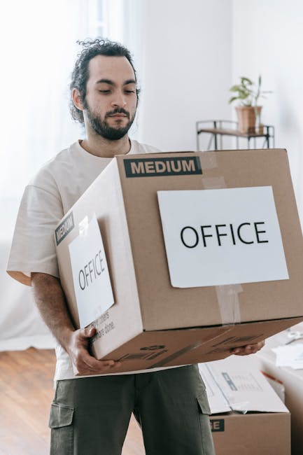 A man with dark hair and a beard, dressed in a beige t-shirt and grey cargo trousers, is indoors carrying a medium-sized cardboard box labeled 'OFFICE' with packing tape. The box appears to contain office equipment or supplies, and has 'MEDIUM' marked on the top left corner. He stands in a well-lit room with light-colored walls and wooden flooring, holding the box with both hands at waist level. In the background, there is a shelf with a potted plant and some decorative items, along with a window allowing natural light into the space. Additional packed boxes are visible on the floor, indicating an ongoing packing or home relocation process. This scene illustrates part of a furniture transport and packing process undertaken by a professional removal service, such as Man and Van Kentish Town, specialising in house removals and office relocation services.