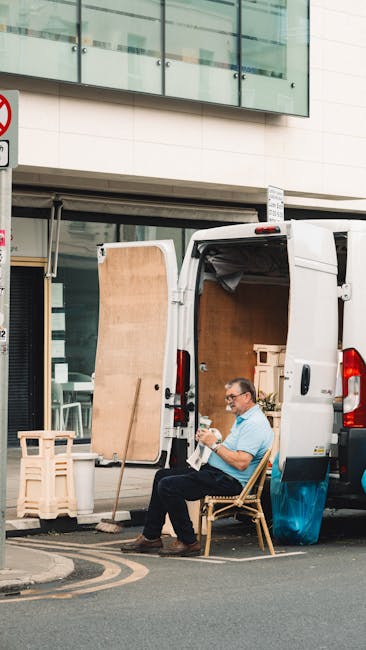 A man wearing glasses and a light blue shirt is seated on a wooden chair next to a white delivery van parked on a city street with modern office buildings in the background. The van’s rear doors are open, revealing an interior loaded with cardboard boxes, wooden furniture, and packing materials such as bubble wrap and plastic wrapping. Leaning against the van are two wooden pallets and a large flat wooden panel, possibly a door or a tabletop, indicating the process of furniture transport and home relocation. The scene appears to be part of a moving or packing operation by Man and Van Kentish Town in the context of house removals for small businesses, with the man taking a break during the loading process of household or office items for a professional relocation service.