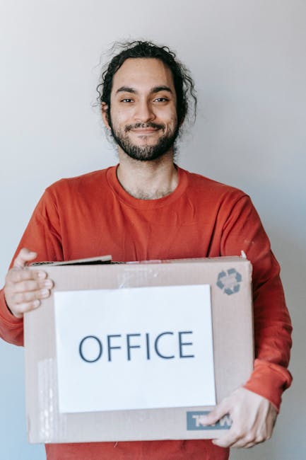 A man with dark hair and a beard, dressed in a beige t-shirt and grey cargo trousers, is indoors carrying a medium-sized cardboard box labeled 'OFFICE' with packing tape. The box appears to contain office equipment or supplies, and has 'MEDIUM' marked on the top left corner. He stands in a well-lit room with light-colored walls and wooden flooring, holding the box with both hands at waist level. In the background, there is a shelf with a potted plant and some decorative items, along with a window allowing natural light into the space. Additional packed boxes are visible on the floor, indicating an ongoing packing or home relocation process. This scene illustrates part of a furniture transport and packing process undertaken by a professional removal service, such as Man and Van Kentish Town, specialising in house removals and office relocation services.