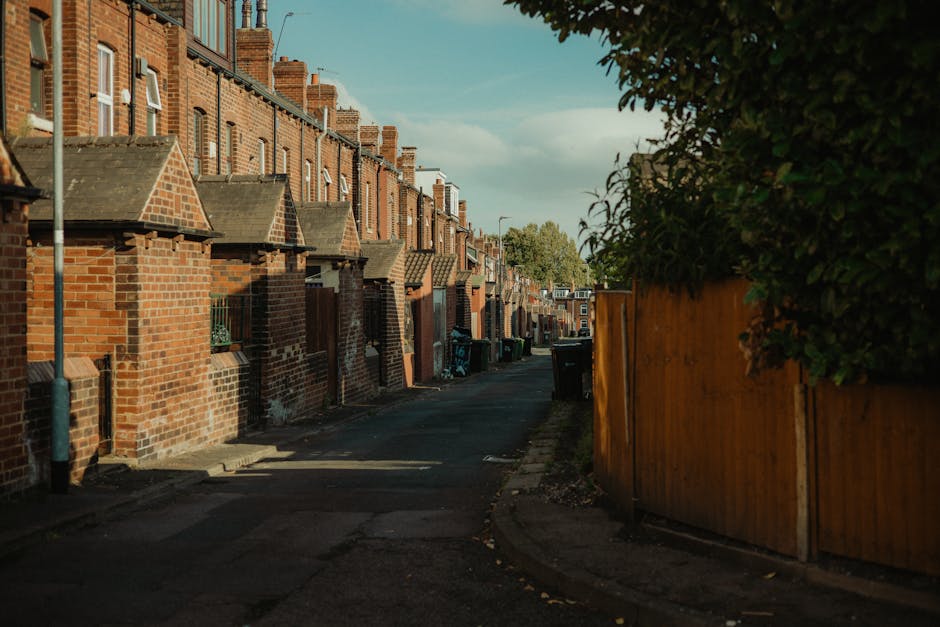 A narrow residential street in Kentish Town lined with row houses constructed from red brick and featuring small front gardens or facades. The houses have pitched roofs and chimneys, with some displaying bay windows. The street is paved with asphalt, with a slight curve leading into the distance, and is shaded by trees and shrubbery on one side, alongside a tall wooden fence. There are black wheelie bins placed along the pavement, and no visible people or vehicles present in the scene. This setting captures a quiet, typical urban environment suitable for house removals or residential relocation activities, with natural daylight illuminating the scene and subtle shadows cast by the houses and trees. As part of moving logistics, the scene suggests a driveway or access point used for loading or unloading household belongings, with no active loading process visible in this image.