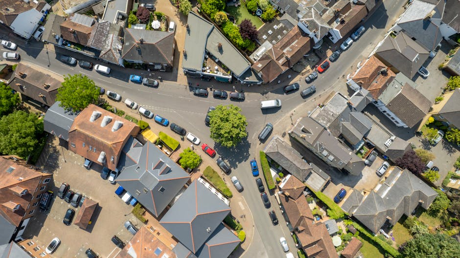 Aerial view of a residential street in Kentish Town with closely spaced houses, some with brick facades and others with tiled roofs, bordered by trees and greenery. Several parked cars line the narrow street, with a white van positioned near the curb, partially visible as it loads furniture and packing boxes wrapped in cardboard, plastic, and fabric blankets. Inside the property, common household items, including a wooden dining table, chairs, and cardboard boxes, are set up for packing and moving, while a household mover from Man and Van Kentish Town appears to be lifting a box and guiding it into or out of the van. The environment is well-lit by natural daylight, with some shadows cast by trees and structures, indicating a typical home relocation process involving furniture transport and packing preparations.