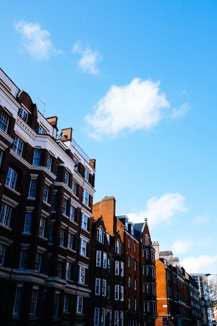 A narrow residential street in Kentish Town lined with row houses constructed from red brick and featuring small front gardens or facades. The houses have pitched roofs and chimneys, with some displaying bay windows. The street is paved with asphalt, with a slight curve leading into the distance, and is shaded by trees and shrubbery on one side, alongside a tall wooden fence. There are black wheelie bins placed along the pavement, and no visible people or vehicles present in the scene. This setting captures a quiet, typical urban environment suitable for house removals or residential relocation activities, with natural daylight illuminating the scene and subtle shadows cast by the houses and trees. As part of moving logistics, the scene suggests a driveway or access point used for loading or unloading household belongings, with no active loading process visible in this image.