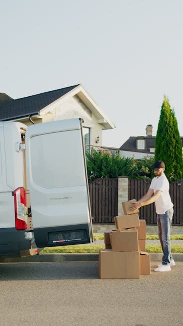 Two men engaged in a home relocation task beside a white van with its rear doors open, filled with cardboard boxes of various sizes packed for moving. The men are lifting a large cardboard box together, with one man wearing a black quilted vest and dark jeans, and the other in a blue jacket with a black padded vest and white sneakers. A black hand truck with two wheels is positioned on the brick driveway nearby, ready for transporting packed items. The scene takes place outdoors, adjacent to a house with a brick wall, a shrub, and trees in the background, under natural daylight, illustrating the process of packing and loading for a furniture transport or house move, with the focus on careful handling and preparation for a move near Kentish Town, as coordinated by [COMPANY_NAME] in their removals services.
