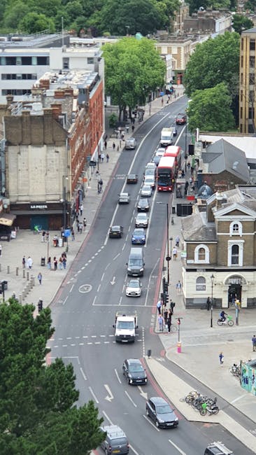 An aerial view of a busy street in Kentish Town lined with trees, with a mix of residential and commercial buildings on either side. Several vehicles, including cars, vans, and a double-decker bus, are moving along the road, while some are parked along the curb. Pedestrians are walking on the sidewalks, and bicycles are parked near a small building on the corner. The scene captures the urban environment during daylight, with green foliage providing contrast to the built surroundings. This setting is indicative of a typical street scene that may be associated with house removals and moving services, emphasizing the importance of efficient furniture transport and loading processes, such as loading vans or trucks from the curbside, possibly coordinated by companies like Man and Van Kentish Town involved in home relocation, packing, and moving logistics.