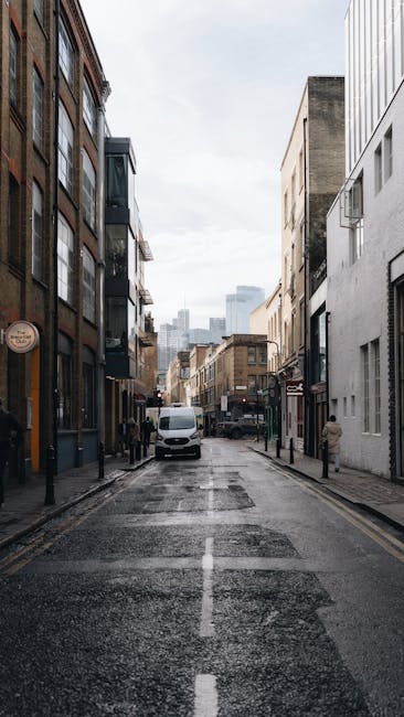 A narrow urban street in Kentish Town with multi-story buildings on both sides, featuring a mix of modern and older architectural styles. The pavement is wet, suggesting recent rain, and there is a white van stationary in the middle of the road, possibly involved in a home relocation or furniture transport. Black bollards line the curb, and pedestrians are visible further down the street. The scene is illuminated by natural daylight under an overcast sky, creating a neutral, professional environment associated with moving and packing services. This setting could relate to a house removal process coordinated by Man and Van Kentish Town, highlighting the typical street environment for loading or unloading furniture and packing materials during a move.